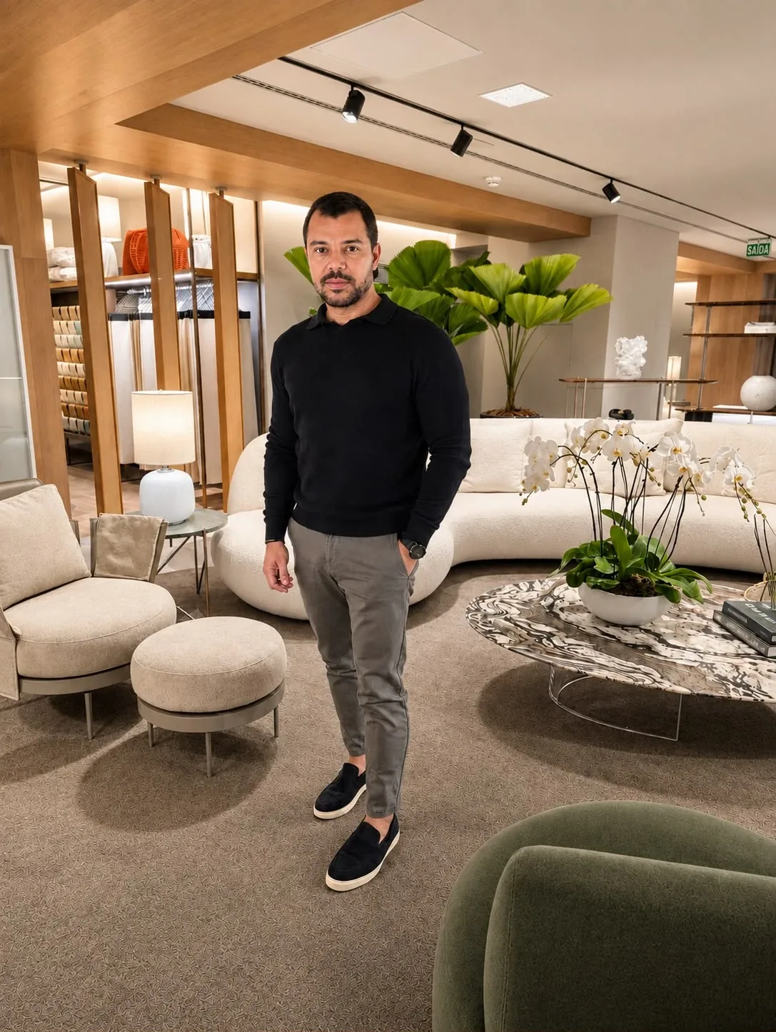 Kenio Carvalho em um showroom de mobiliário, entre sofás curvos e orquídeas